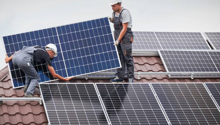 An image of a solar panel installation on the roof of a commercial building in Summerville, SC. The installation team can be seen working on the roof, positioning the panels to capture maximum sunlight. The city skyline can be seen in the background, with clear blue skies and a few clouds. Tools, equipment, and safety gear are scattered on the roof.